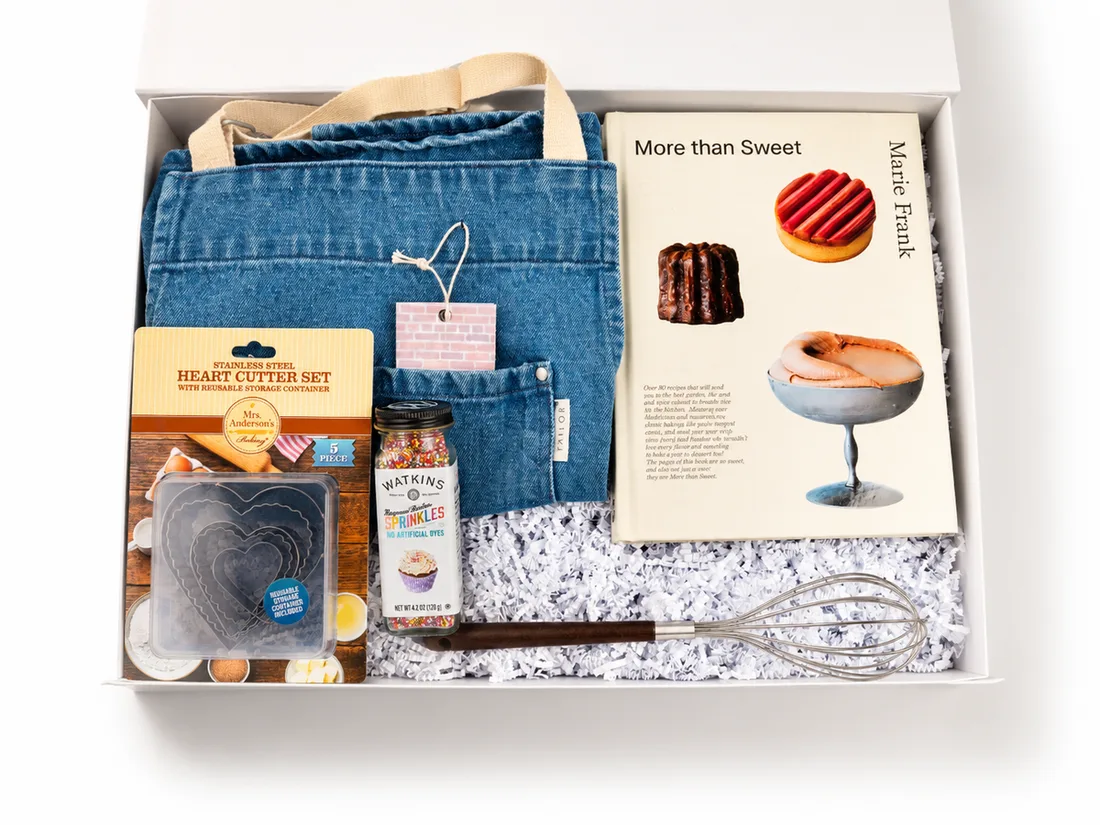 White gift box holding a denim apron, a baking cookbook, cutter set, sprinkles and a whisk